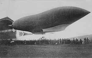 An airship hangar at lower/mid left; an airship with a pointed bow, which has just left the hangar, stretches from mid left to mid right